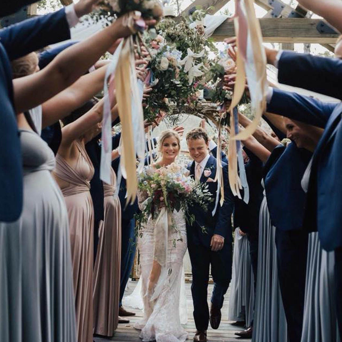 Newlyweds walk through an archway of raised arms holding ribbons and bouquets, flanked by wedding party.