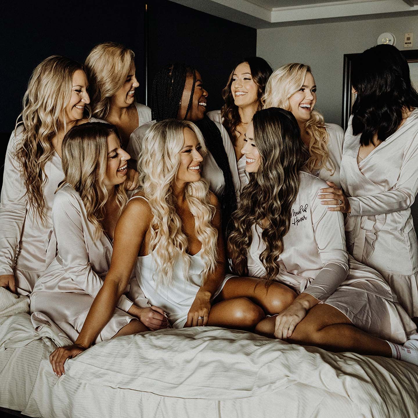 Bridesmaids in silky robes on a bed, smiling and laughing.