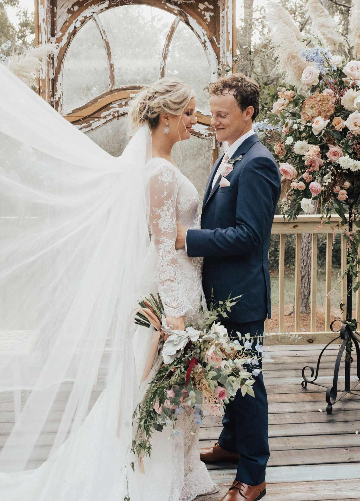 Bride and groom embrace on wooden deck, long veil blowing, surrounded by flowers.