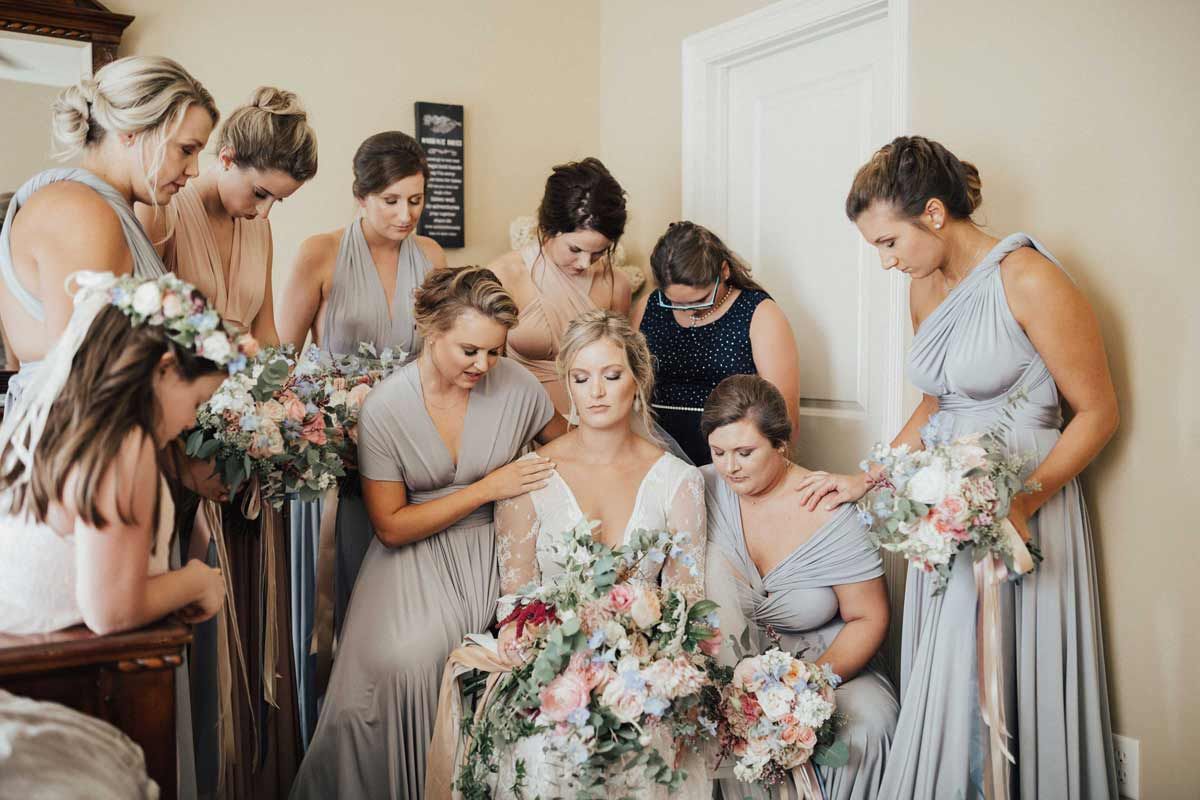 Bride surrounded by bridesmaids, seated, praying, with bouquets in soft-colored dresses.