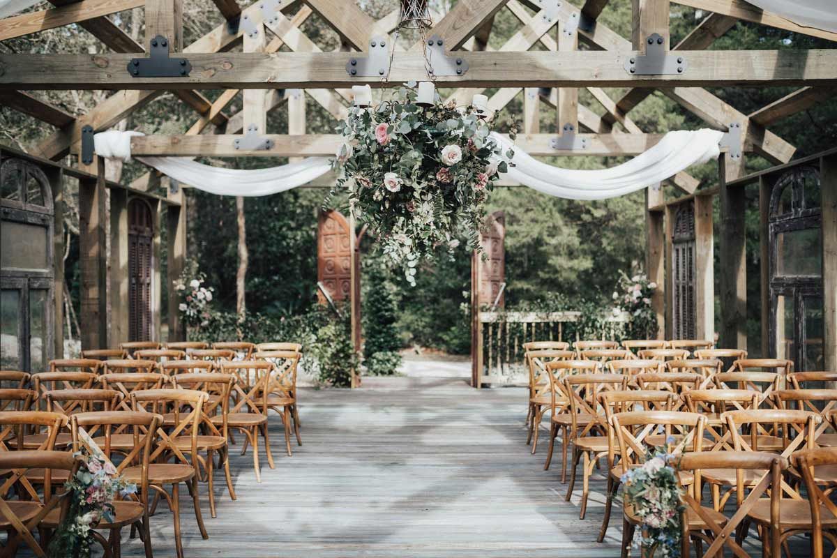 Wooden-framed outdoor wedding ceremony setup. Rows of wooden chairs face an arbor with floral arrangement and draped fabric.