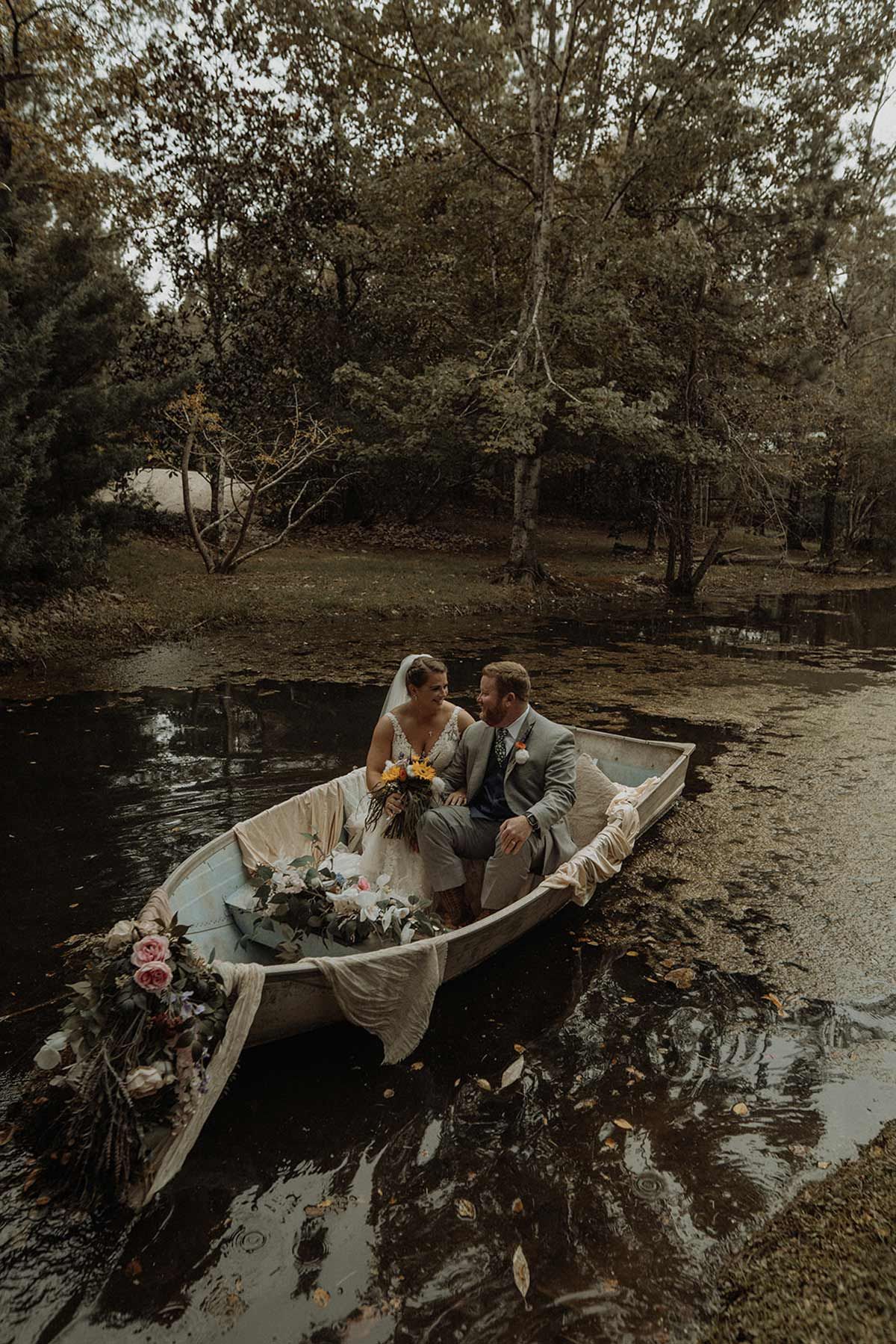 Couple in wedding attire on a boat in a pond, decorated with flowers.