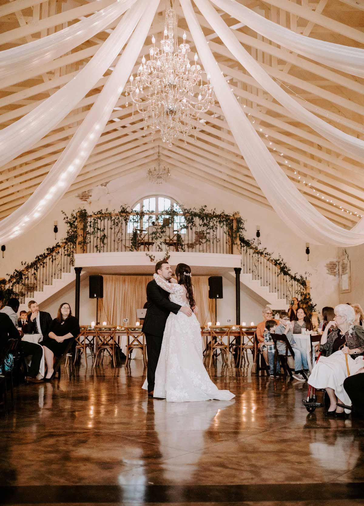 Couple dancing in a wedding reception hall, draped with white fabric, and a chandelier overhead.