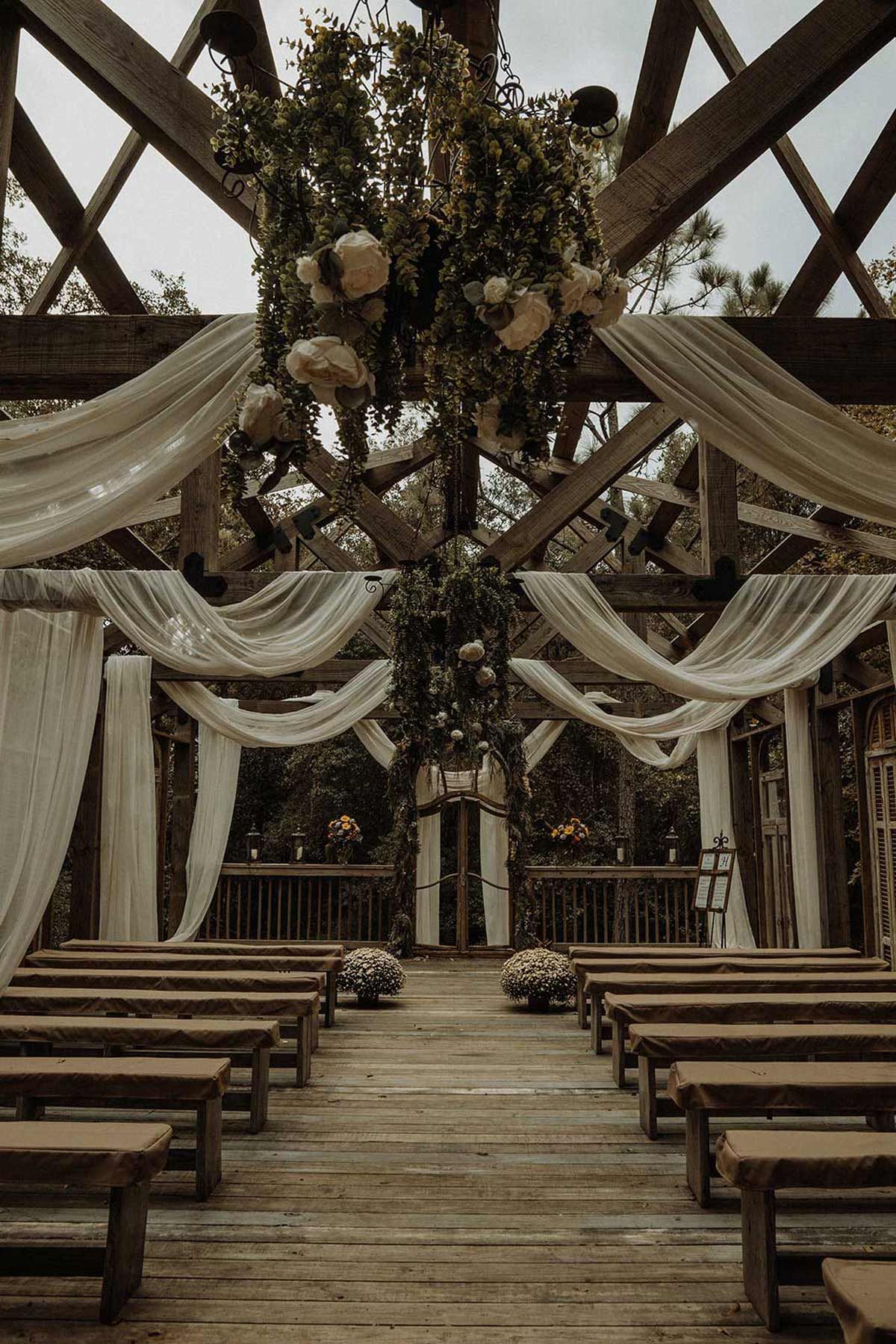 Wedding ceremony setup under a wooden pergola with draped fabric and floral arrangements.
