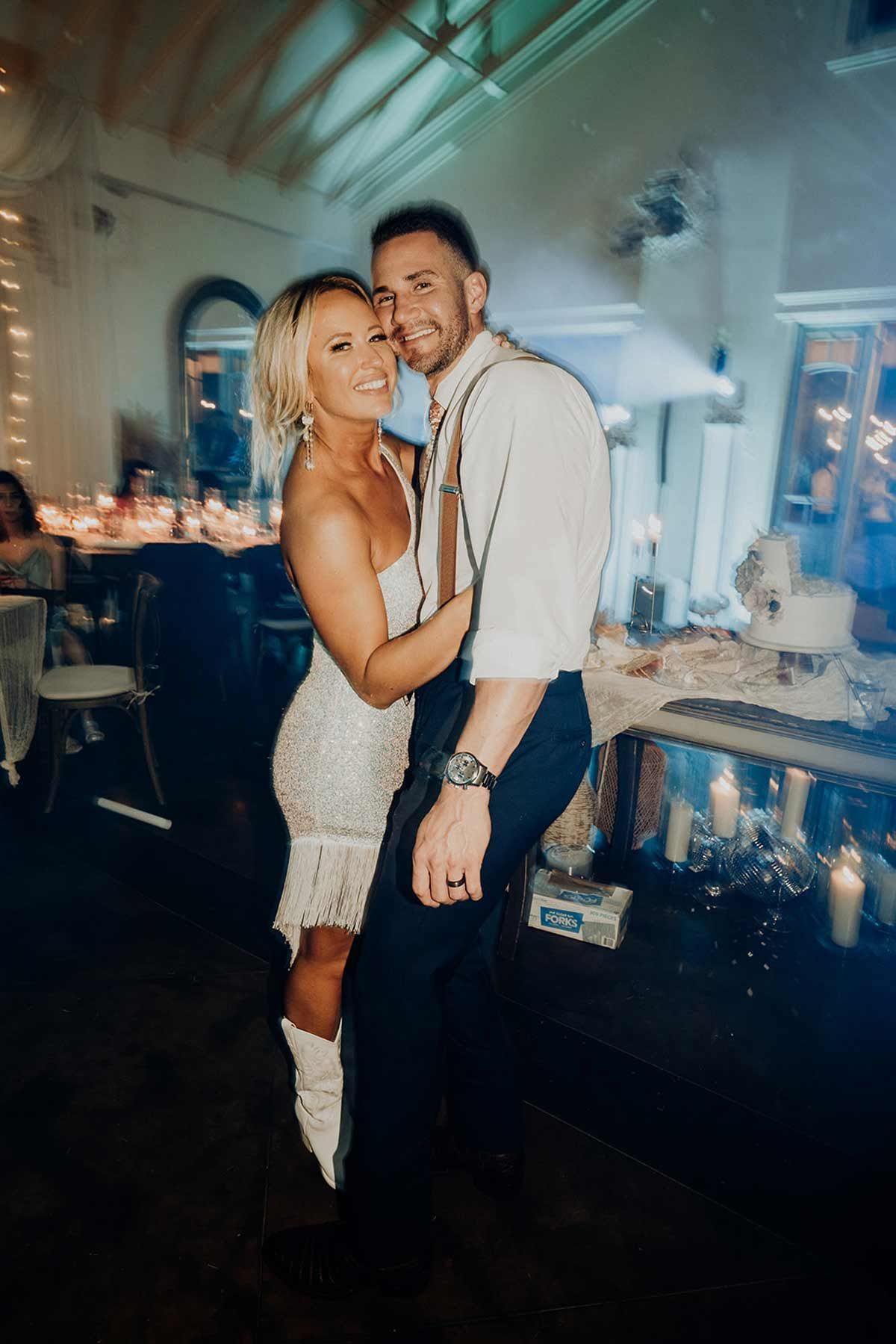 Couple dancing close together at a reception; woman in white dress, man in suspenders, both smiling.