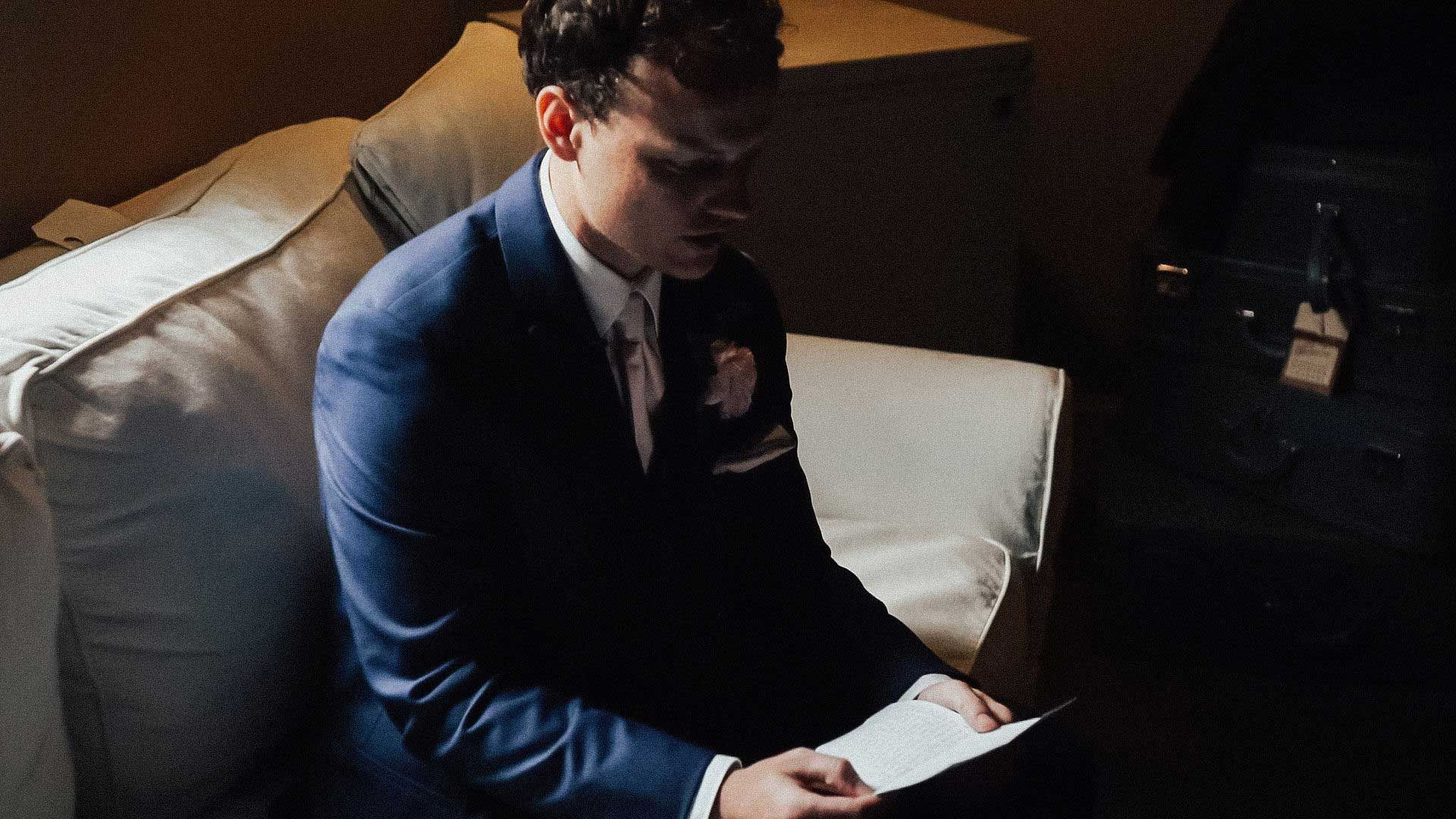 Man in a blue suit seated, reading a letter in a dimly lit room.