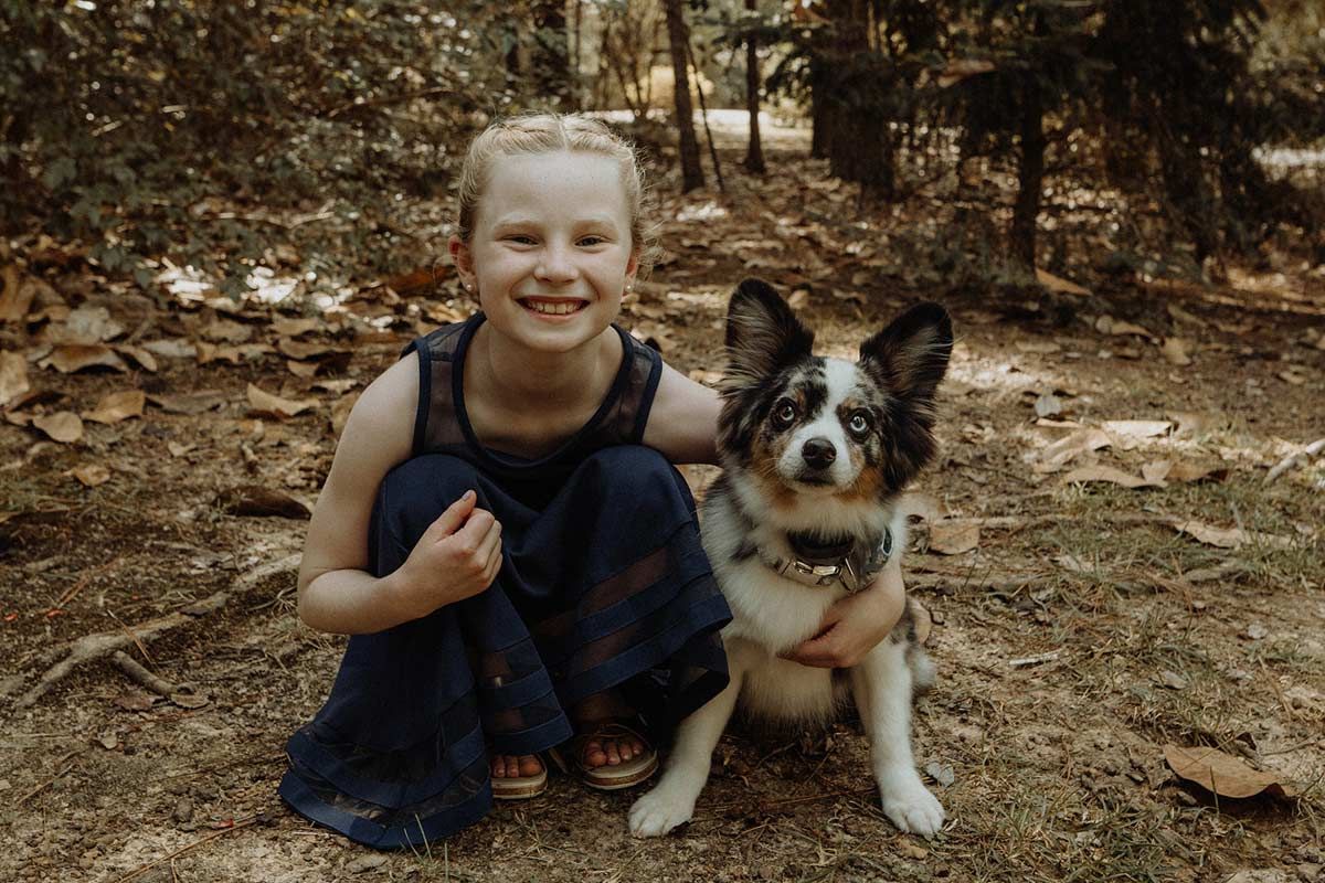 Girl with blonde hair smiles, crouching next to a dog with blue eyes and a speckled coat outdoors.