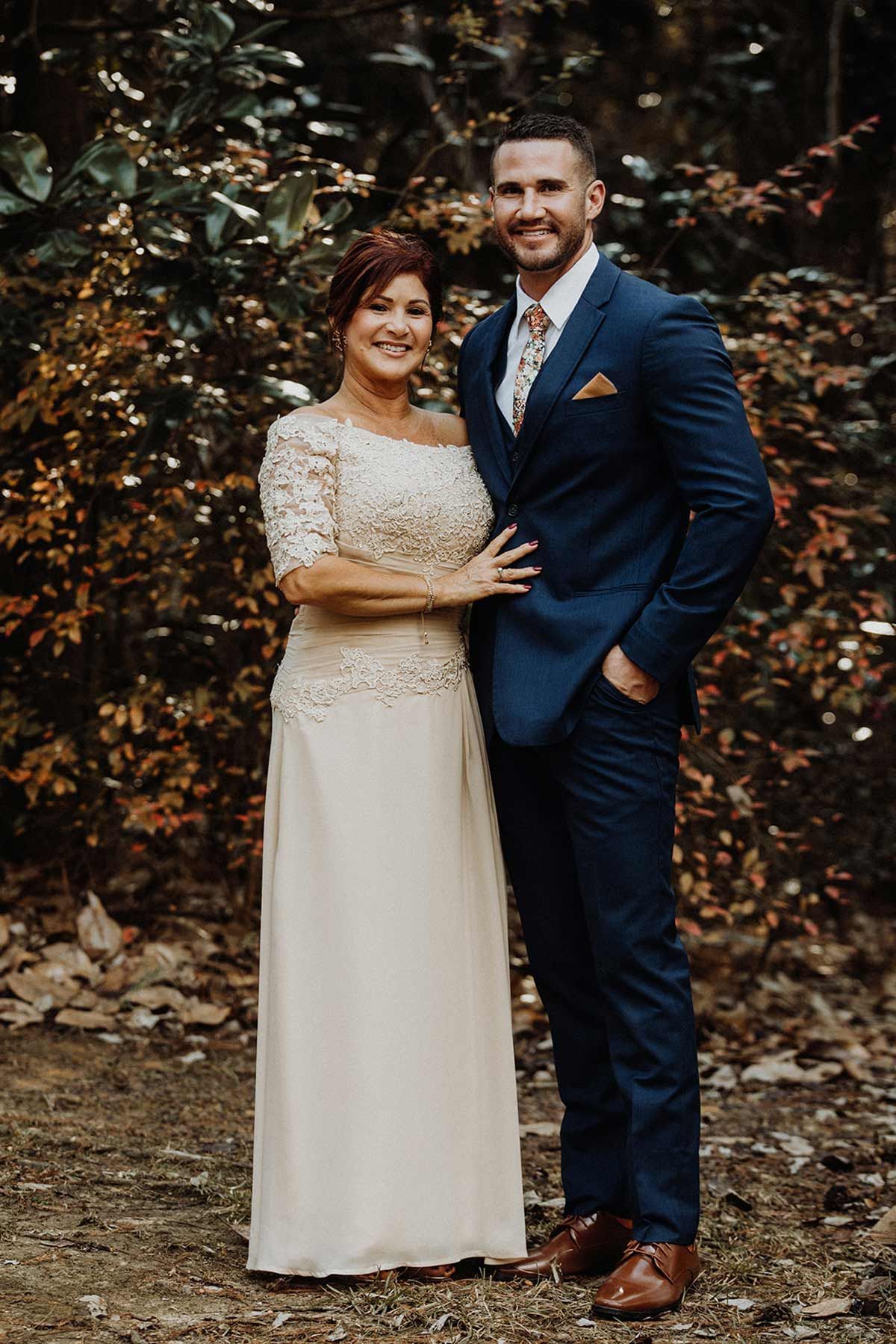 Couple in formal wear standing outside, woman in cream dress with lace, man in blue suit.