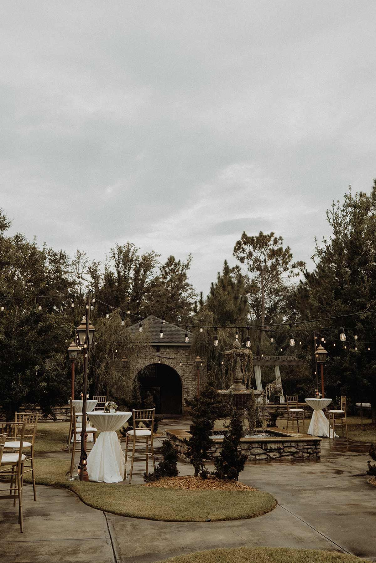 Outdoor event space with stone fountain, archway, and white-covered tables under a cloudy sky.