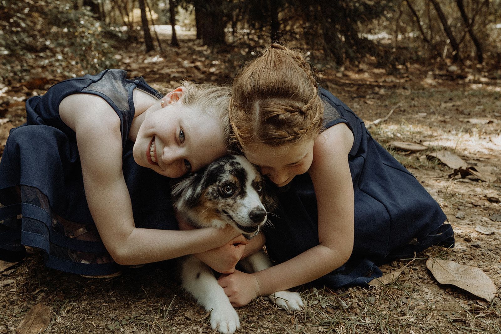 Two girls in blue dresses hug a dog outdoors.