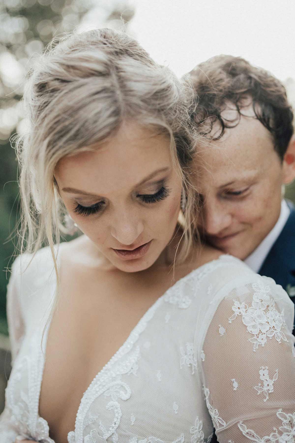 Bride in wedding dress looks down, groom hugs her.