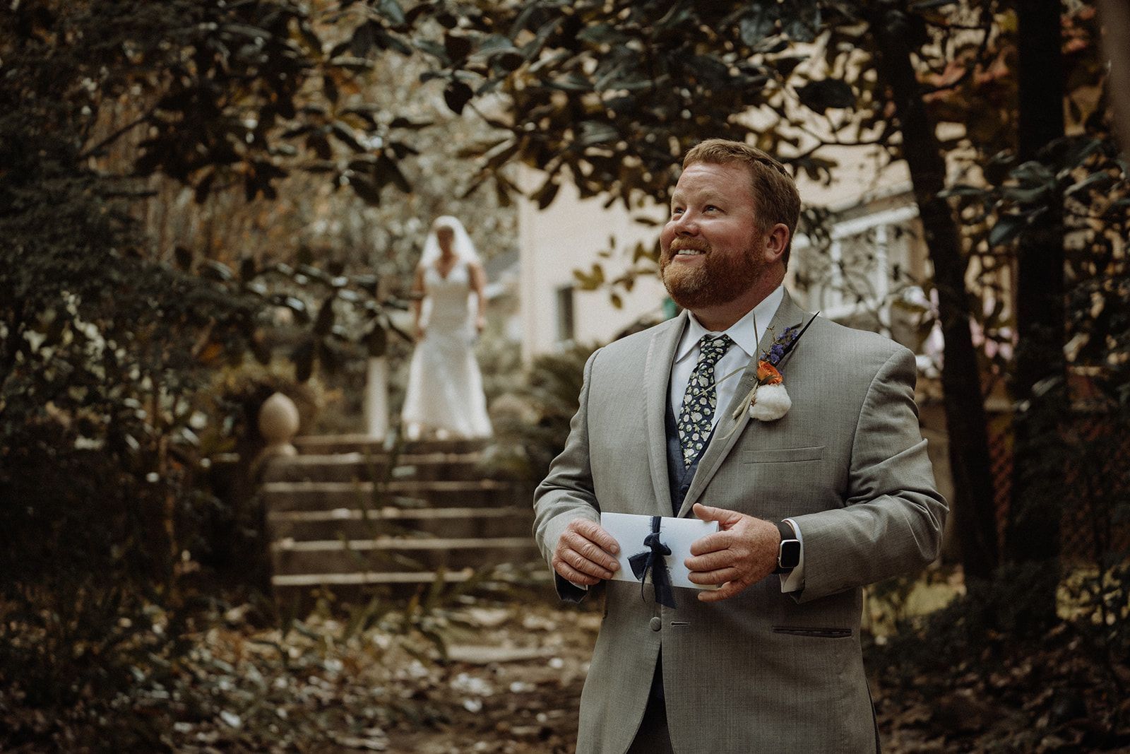 Groom smiles, holding a card, as bride walks towards him up outdoor stairs. Wedding day.
