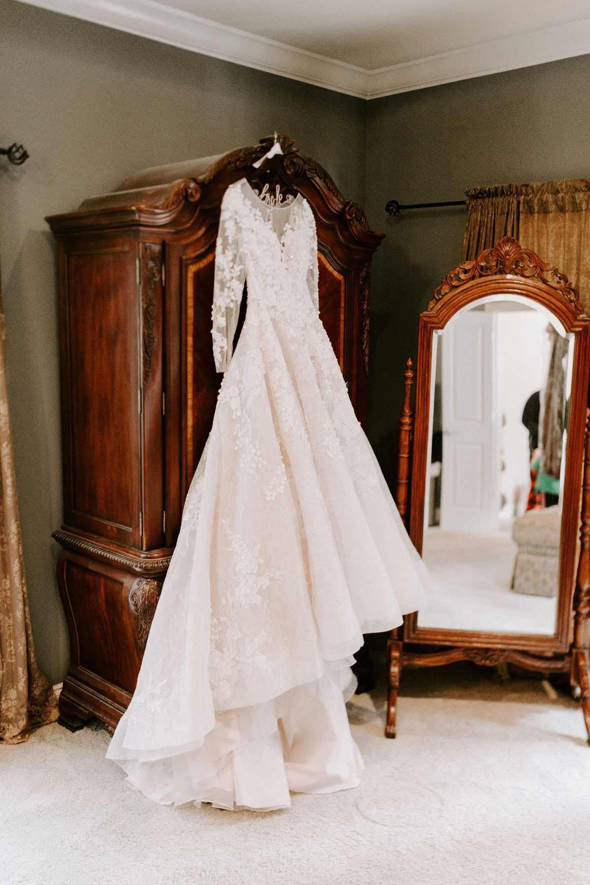 Wedding dress hanging on a hanger inside a room with a wooden wardrobe and mirror.