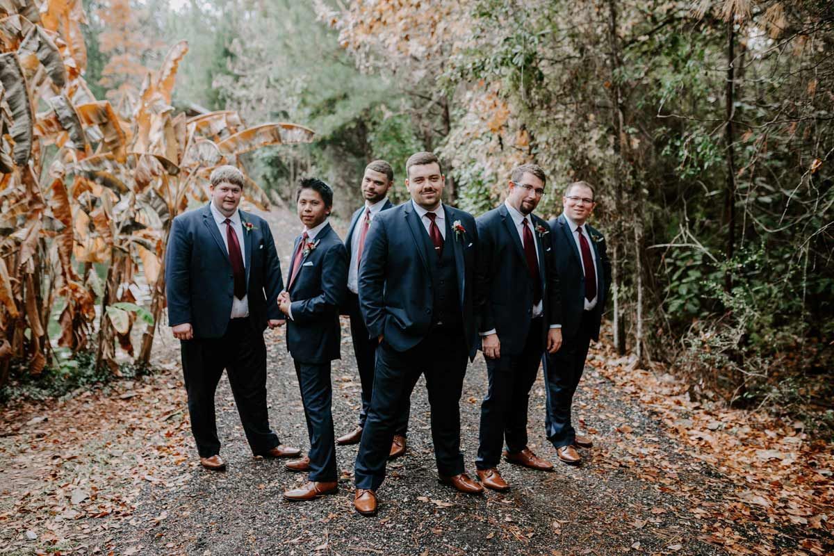 Groomsmen in navy suits with red ties stand on a path in a wooded area.