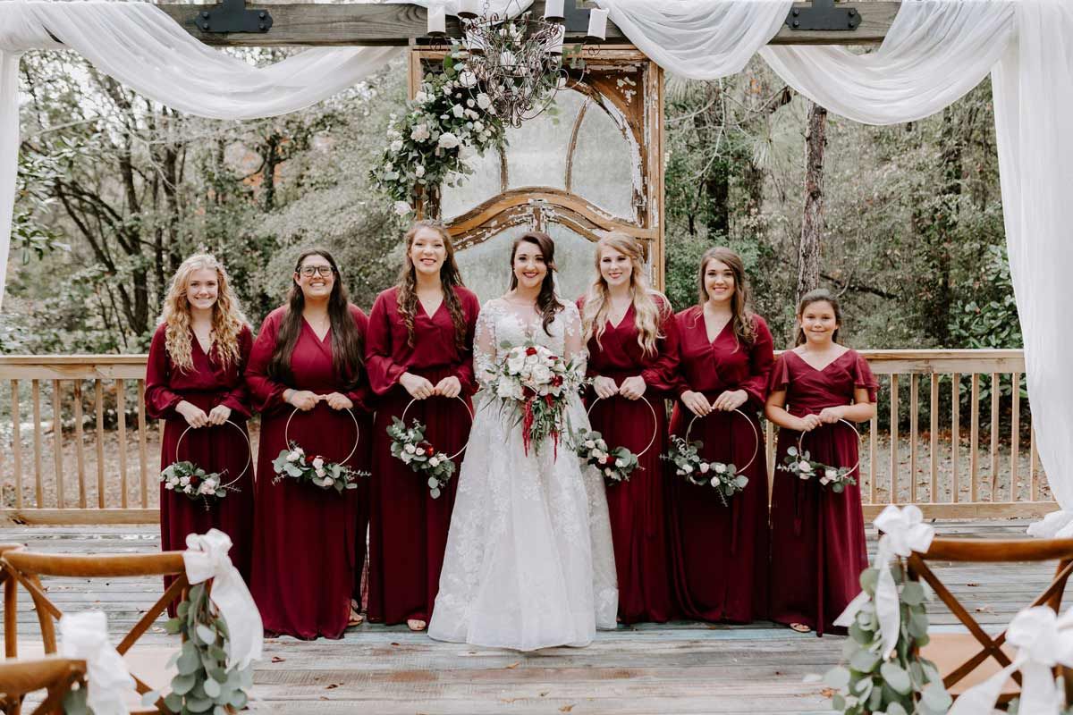 Bride and bridesmaids in burgundy dresses posing outdoors, holding wreaths, under a floral arch.