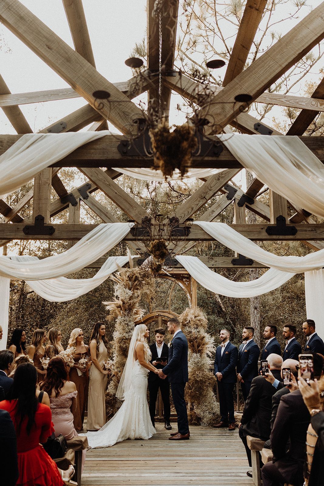 Wedding ceremony: Bride and groom hold hands under a draped wooden arbor; guests watch.