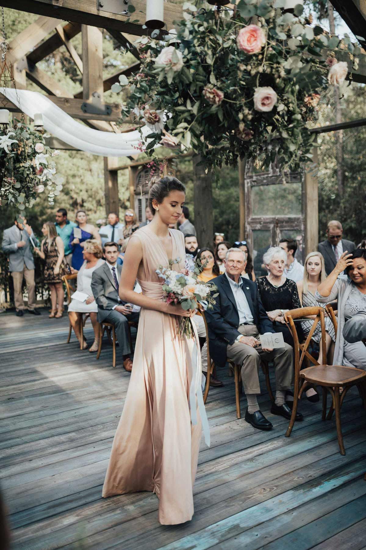 Bridesmaid in a peach dress walks down aisle at outdoor wedding under floral arch. Guests watch.