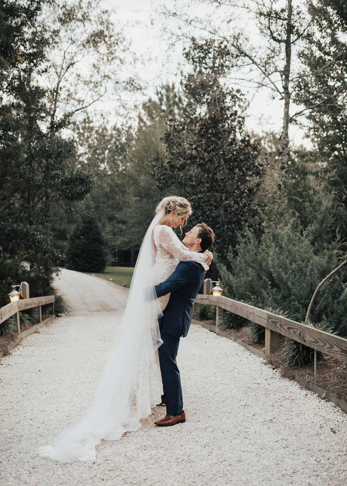 Groom lifts bride in wedding attire on a gravel path, surrounded by trees.