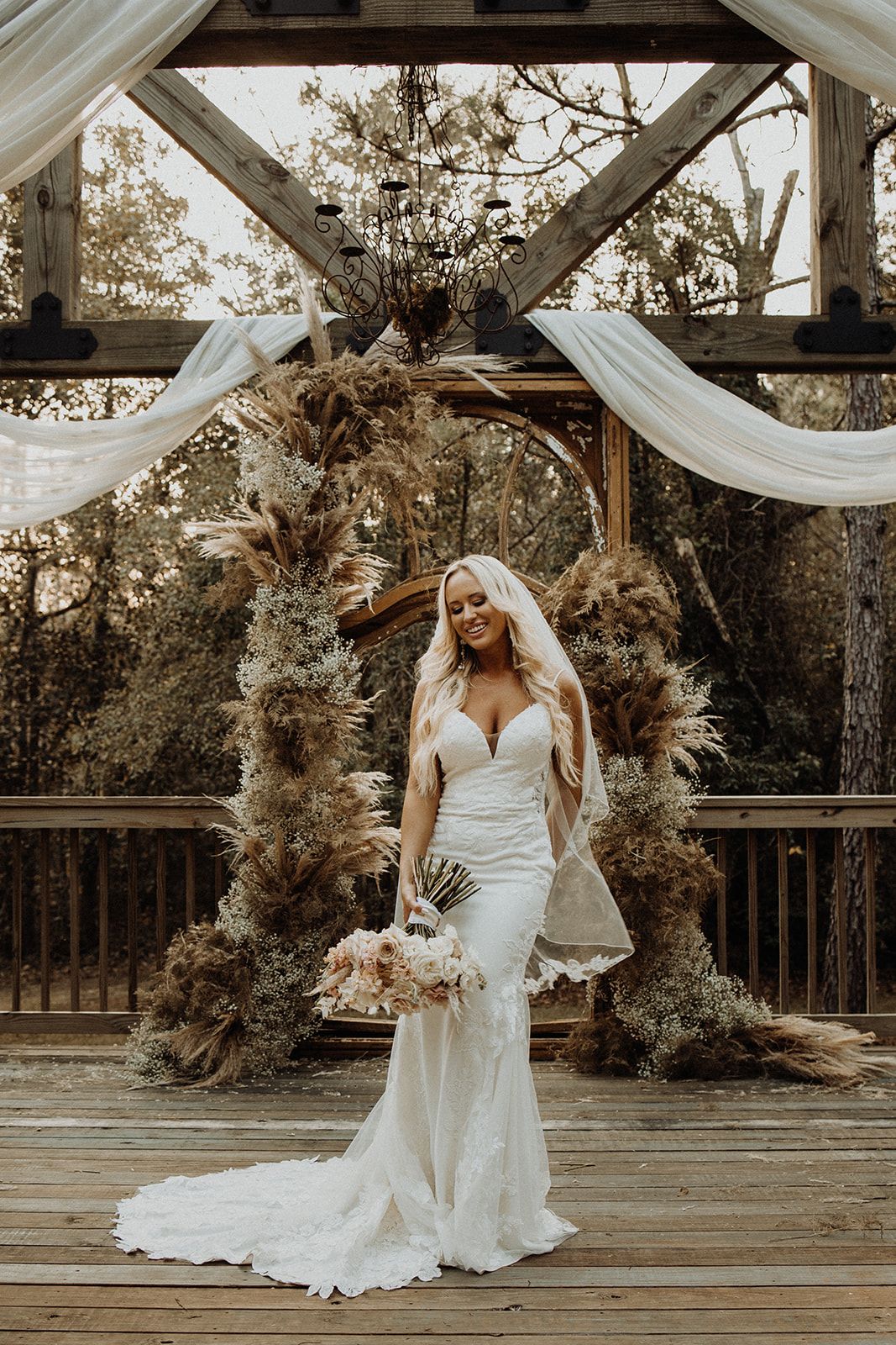 Bride in lace wedding dress smiling, holding bouquet, in front of a decorated wooden arch.