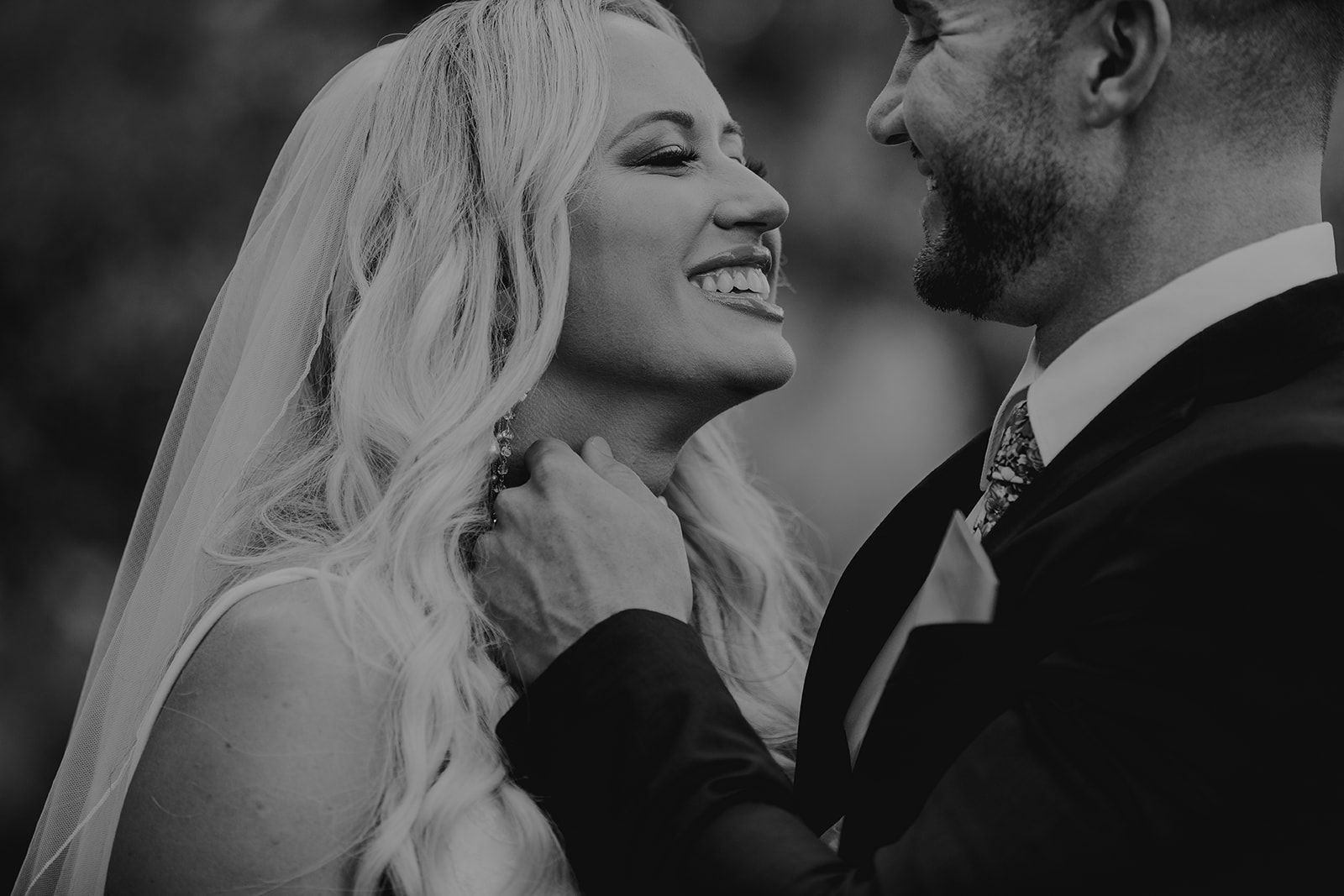 Bride and groom smiling at each other, outdoors. Man's hand on woman's neck, woman in veil. Black and white.