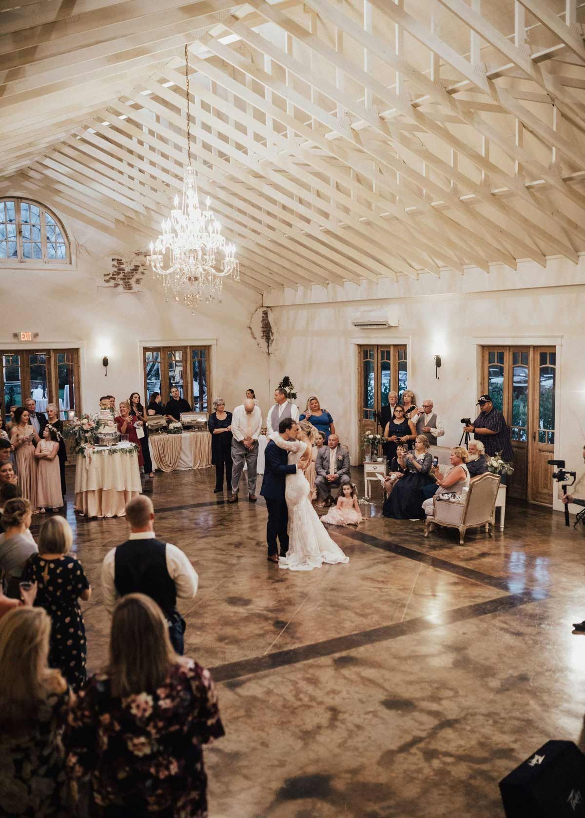 Couple dances at wedding reception; guests watch. Rustic venue with chandelier and exposed beams.