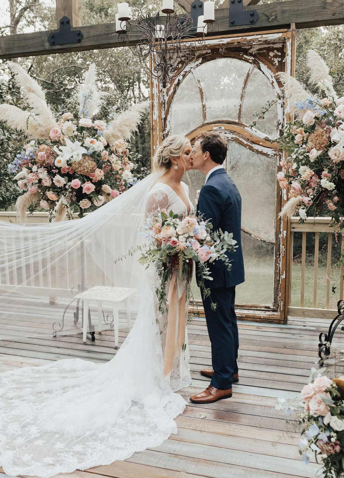 Bride and groom kissing at outdoor wedding ceremony; floral arch, veil, bouquet, wood deck.