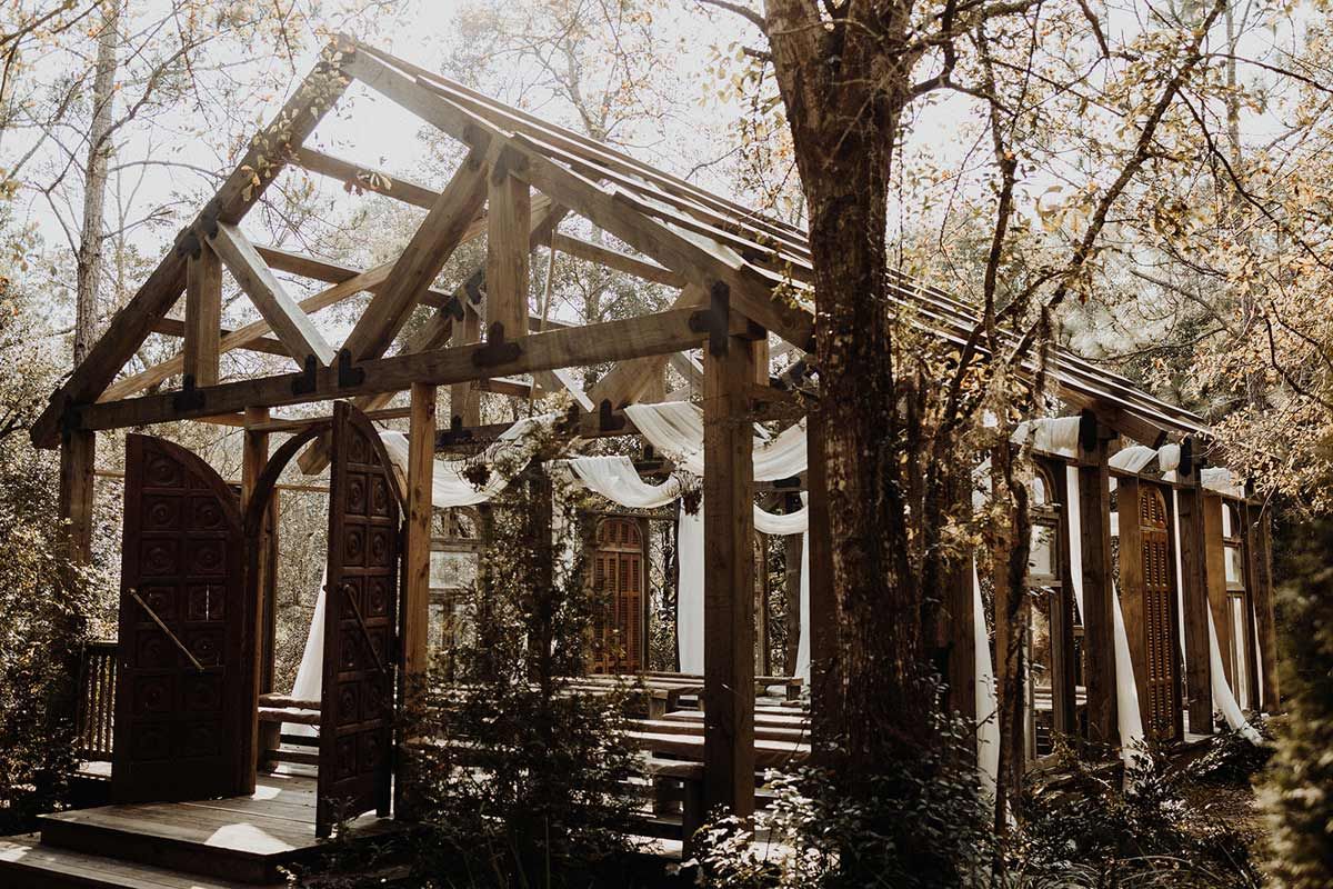 Wooden outdoor wedding pavilion in a forest setting, draped with white fabric, with an open door.