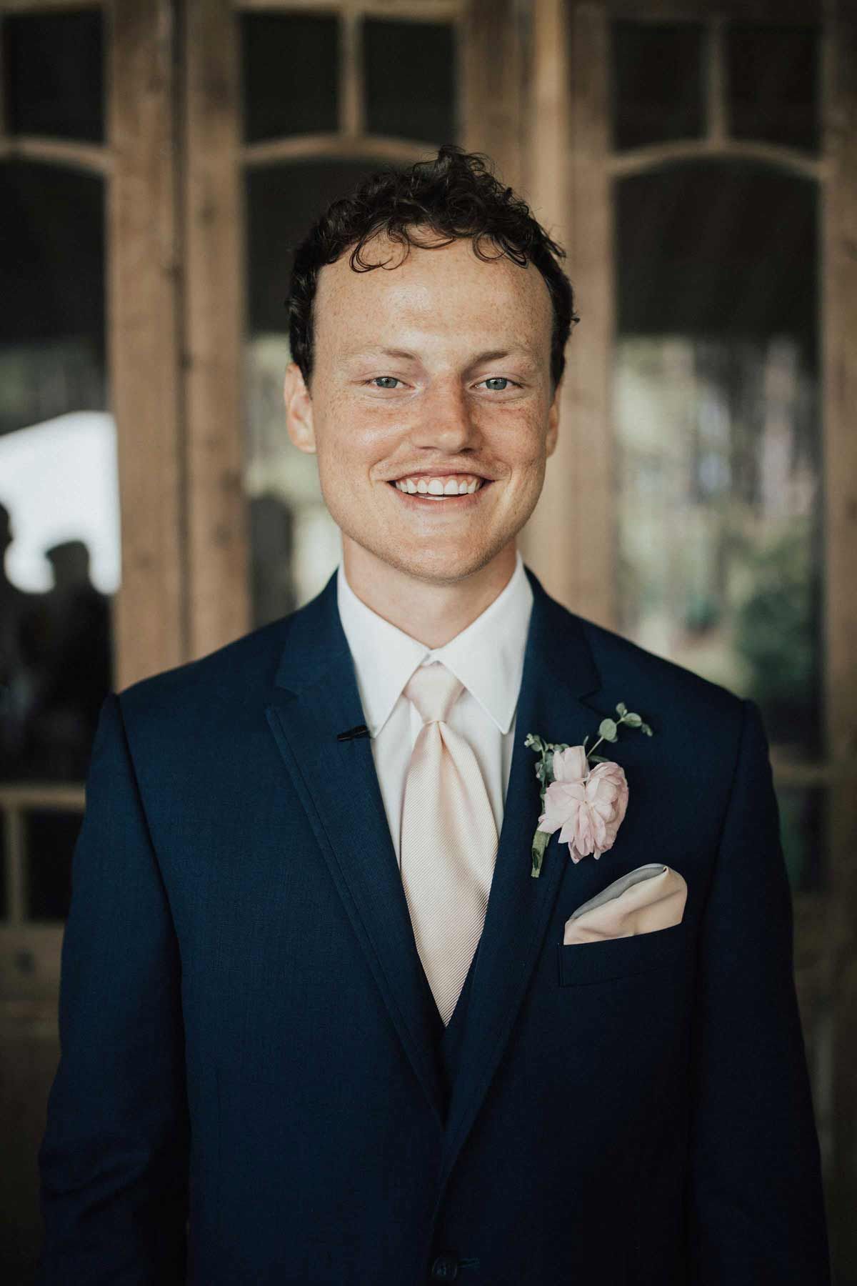 Man in navy suit, blush tie, smiling, flower in lapel, standing outdoors.