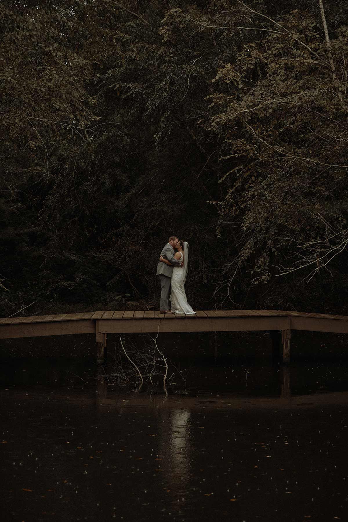 Couple kissing on a wooden dock over water; trees in background.