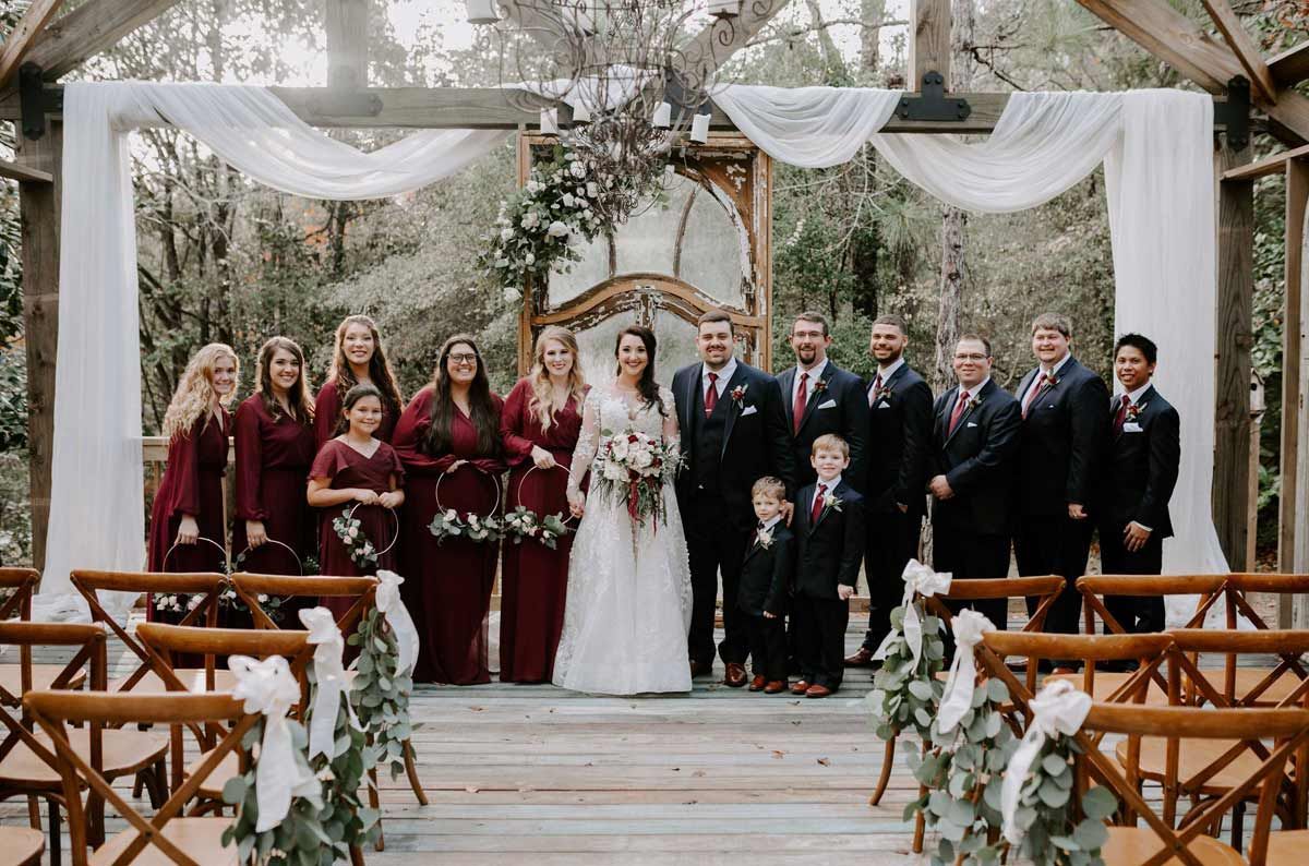Wedding party poses in front of an outdoor archway with draping fabric and floral accents.