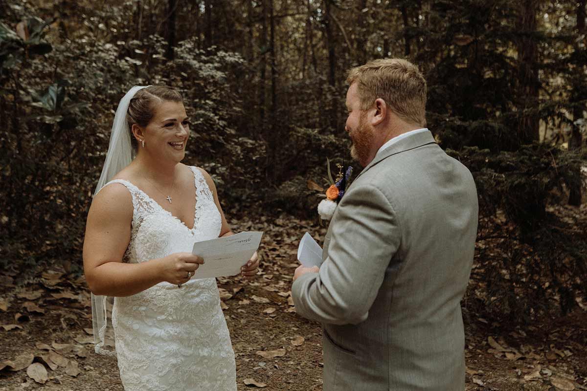 Bride and groom reading letters to each other outdoors in a forest. She wears a white dress, he a gray suit.