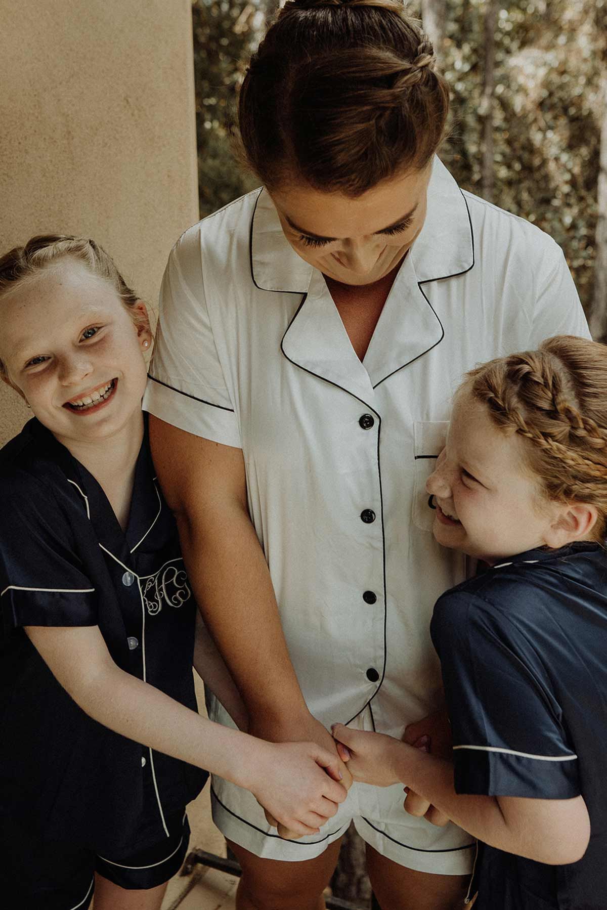 Woman in white pajamas with two children in navy pajamas, all smiling, holding hands.