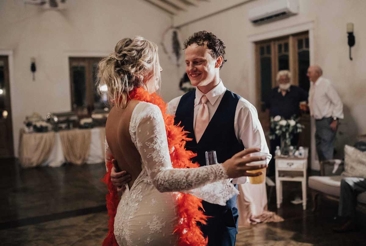 Couple dancing at a wedding reception, woman in white dress, red boa. Man in vest, holding drink.
