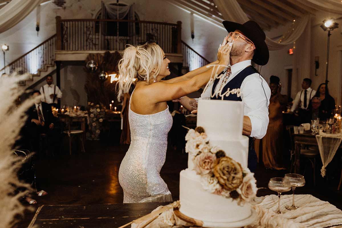 Bride smashes cake on groom's face at wedding reception. Indoor, tiered cake with floral accents.