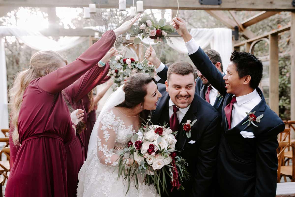 Wedding party celebrates under an arbor. Bride and groom surrounded by bridesmaids and groomsmen with raised arms.