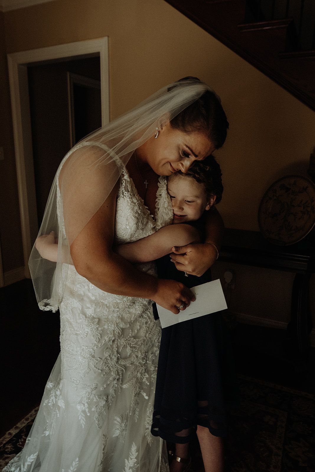Bride in a white dress hugs a child in a dark dress indoors. Both are emotional.
