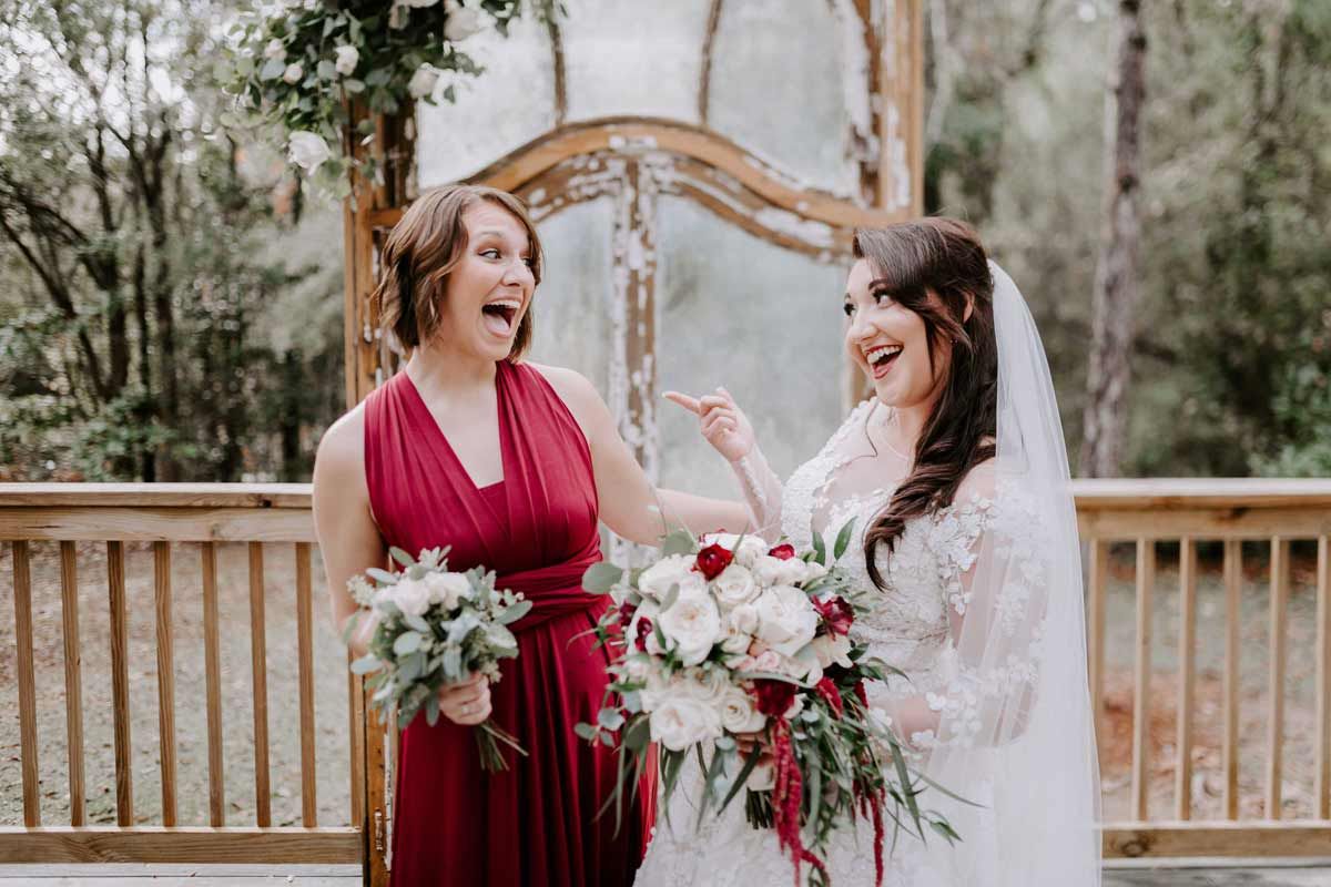 Bride and bridesmaid laughing on a wooden deck. Bride wears white dress with veil, bridesmaid in a red dress. Both hold bouquets.