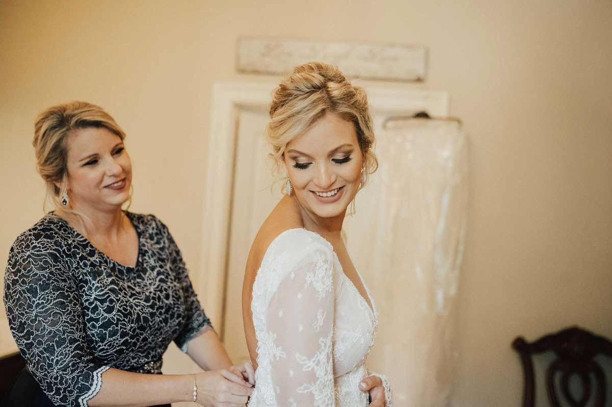 Woman in wedding dress, smiling, as another woman zips the back. Indoor setting with neutral tones.