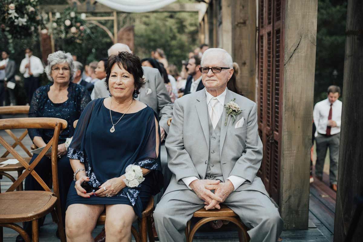 Guests seated at a wedding, waiting. Woman in blue dress and man in gray suit. Outdoors, daytime.