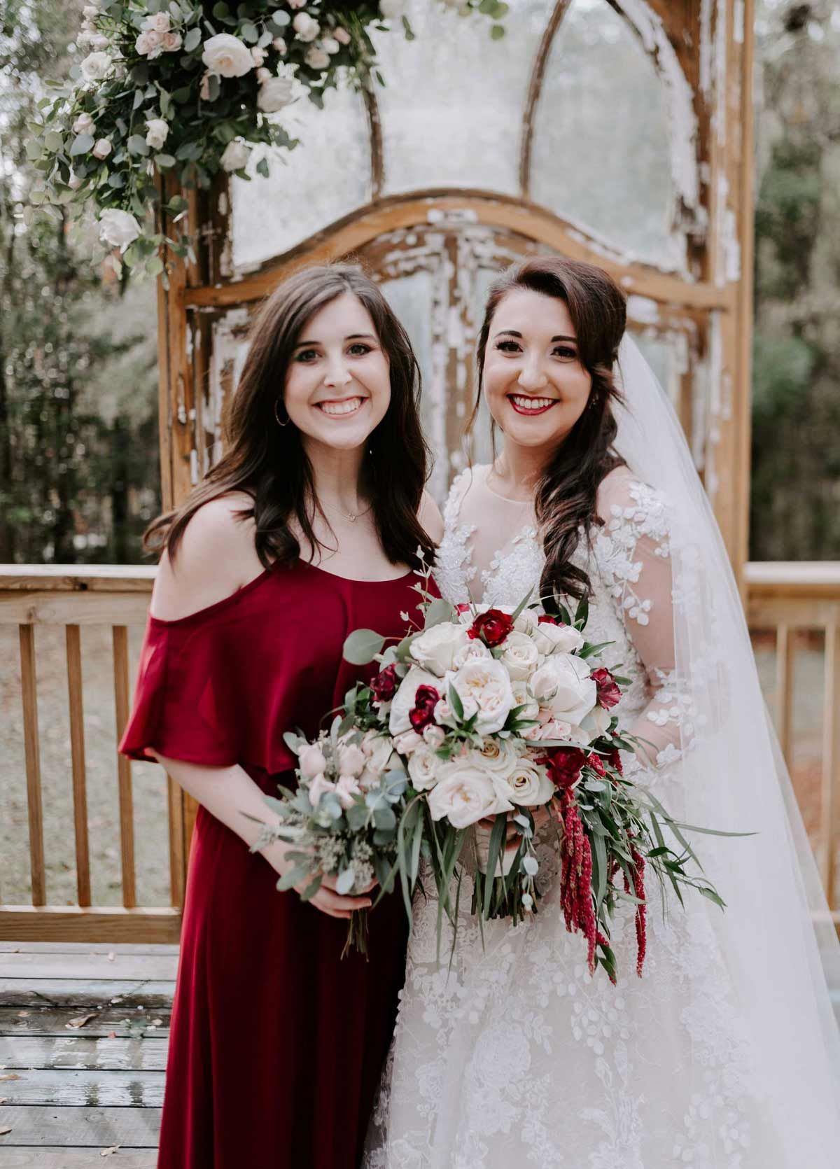 Woman in burgundy dress with bride in a white wedding gown, holding bouquets, smiling outdoors.