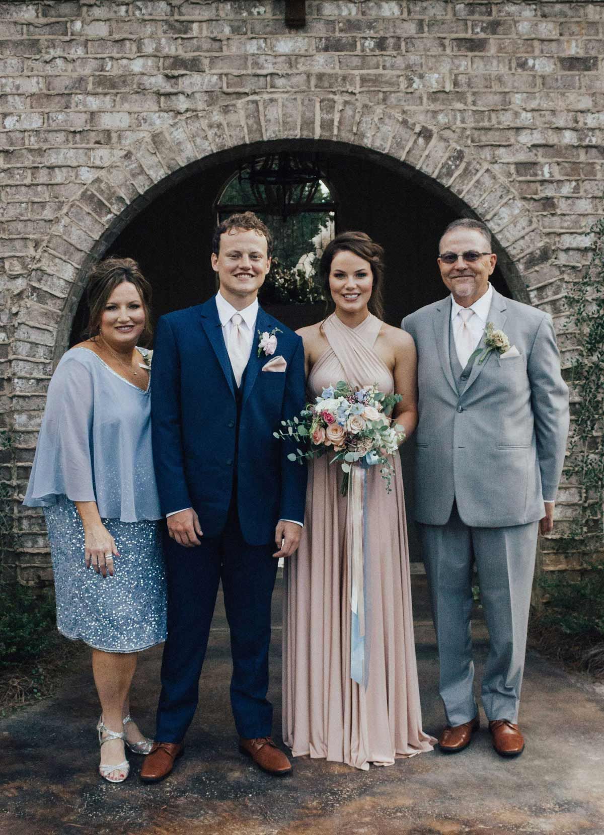 Wedding portrait: couple, parents pose under brick archway. Bride in pink, groom in navy suit.