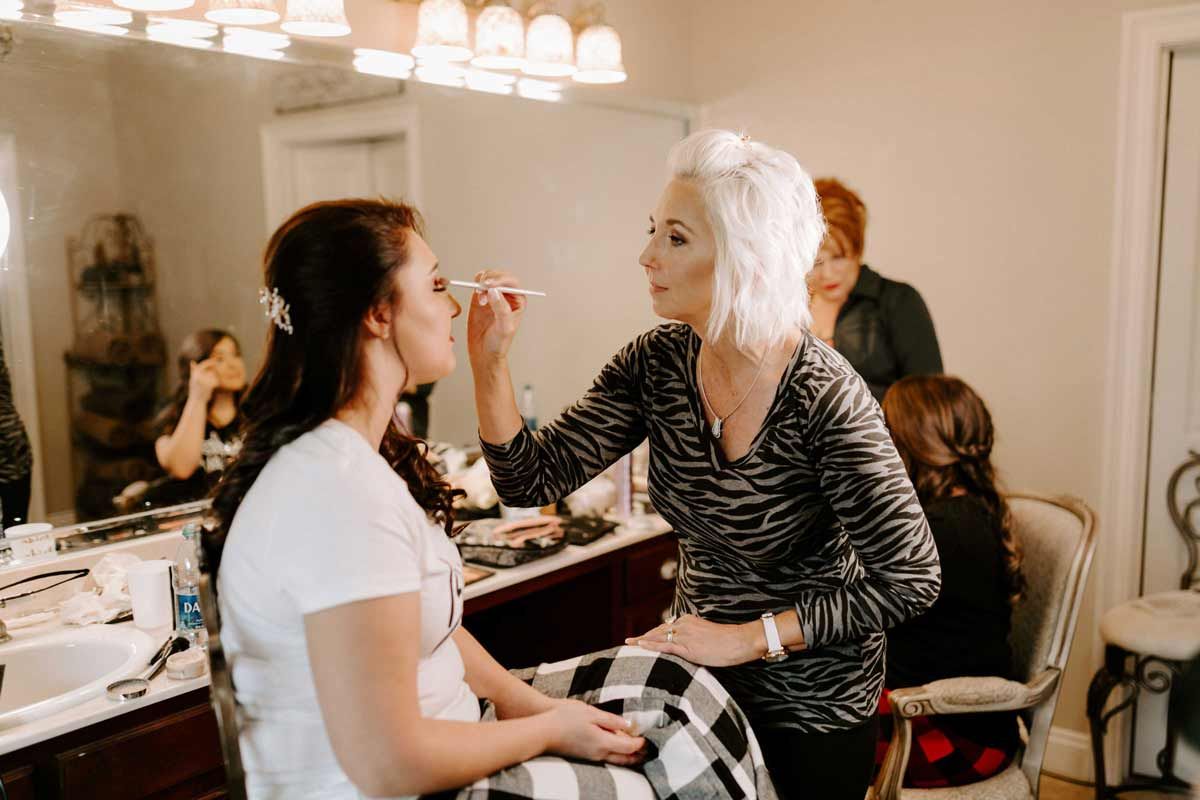 Makeup artist applying makeup to a person seated in front of a mirror in a well-lit room.