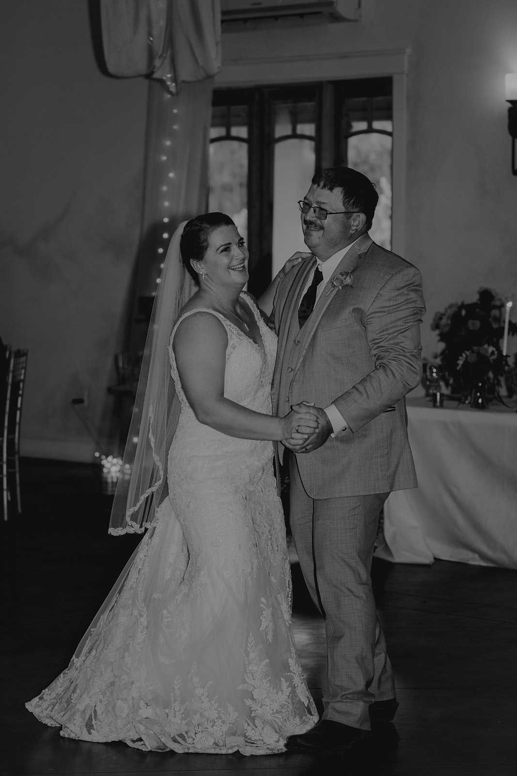 Bride and her father dance at wedding reception, smiles, holding hands, elegant gown, soft lighting.