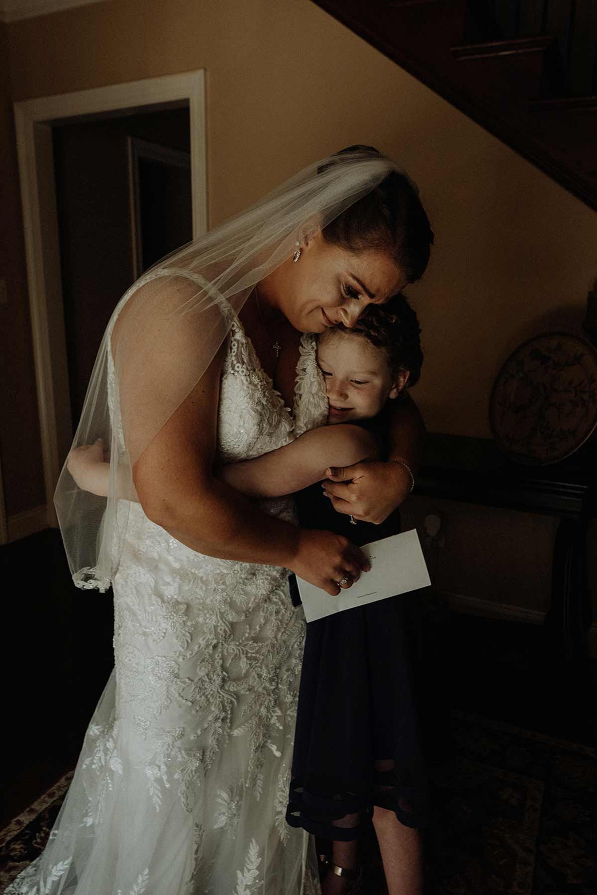 Bride in wedding dress hugs a child in a dark dress indoors. She wears a veil and has her head resting on the child's.