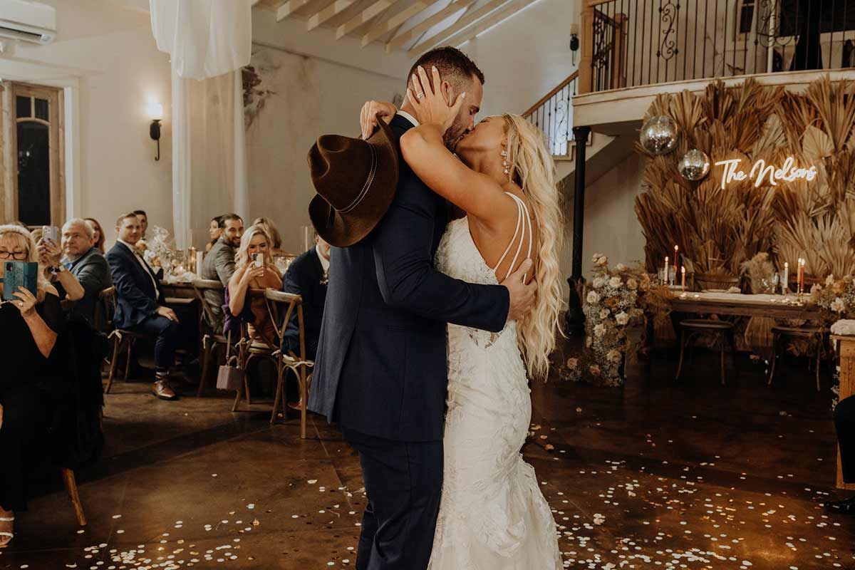 Newlyweds share a kiss during their wedding dance, cowboy hat in hand, guests watch.