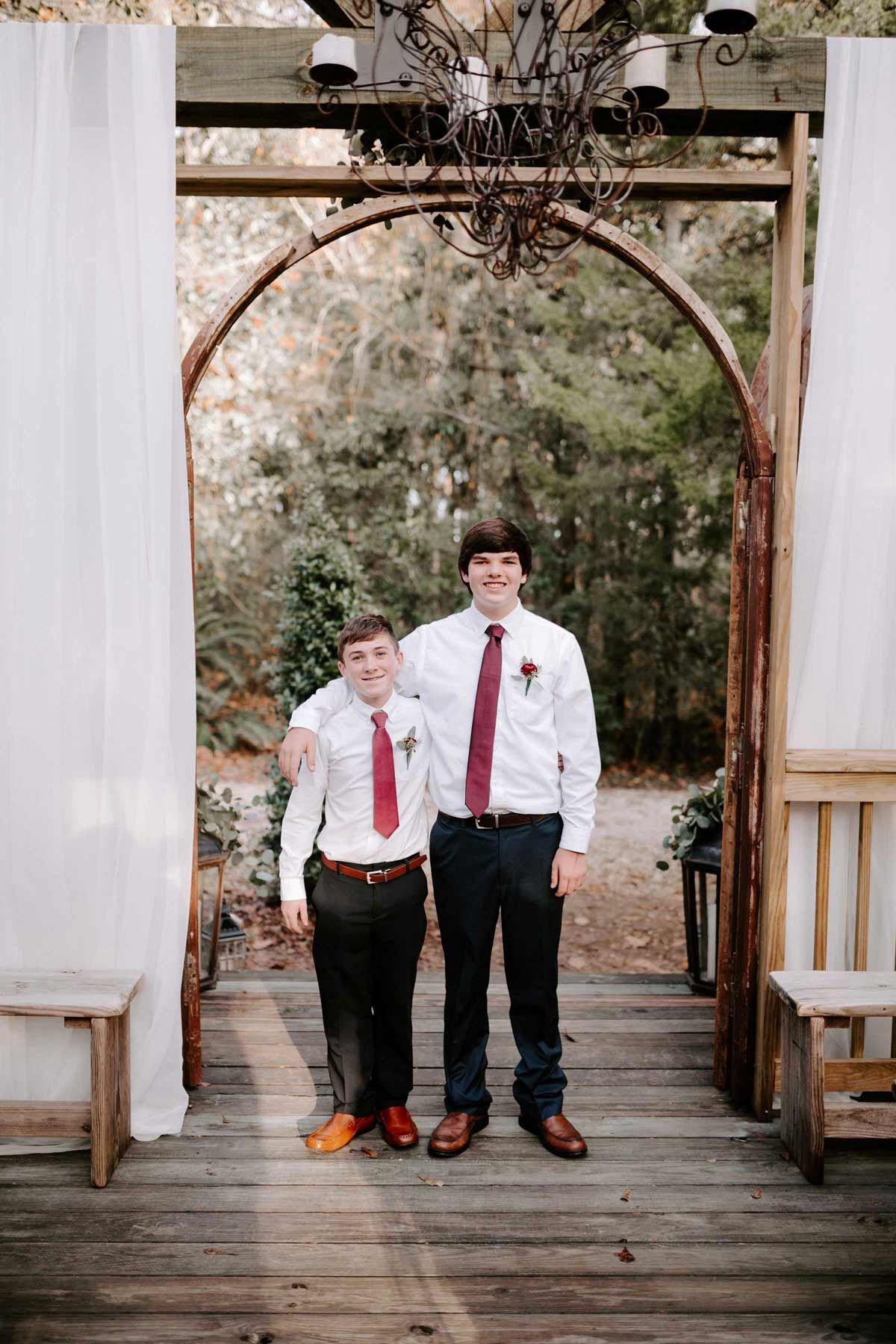 Two people in formal attire stand under an archway, one with an arm around the other's shoulder; red ties, white shirts.