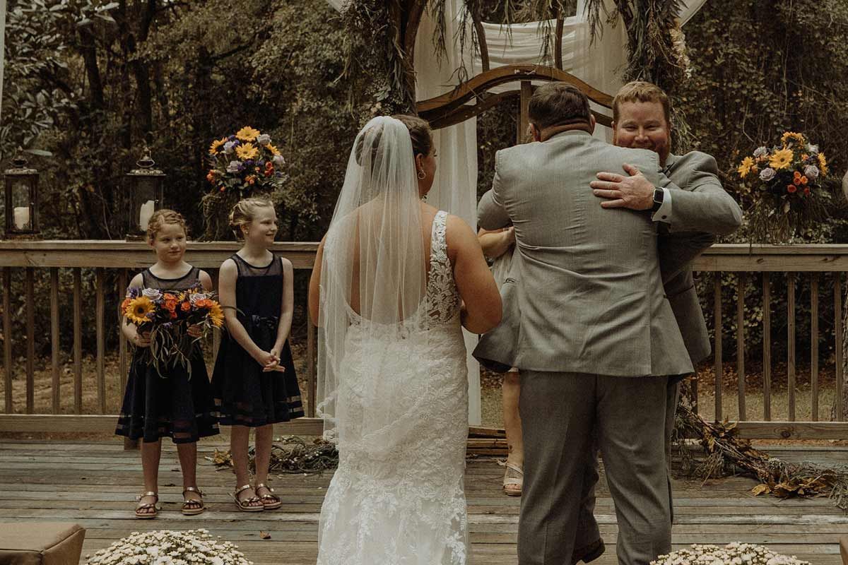 Bride and groom hugging at an outdoor wedding ceremony. Two young girls stand nearby.