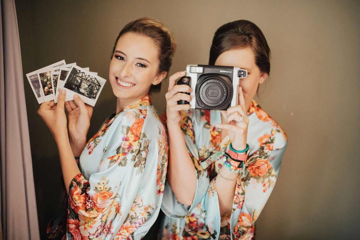 Two women in floral robes, one holding instant photos, the other with an instant camera, smiling.