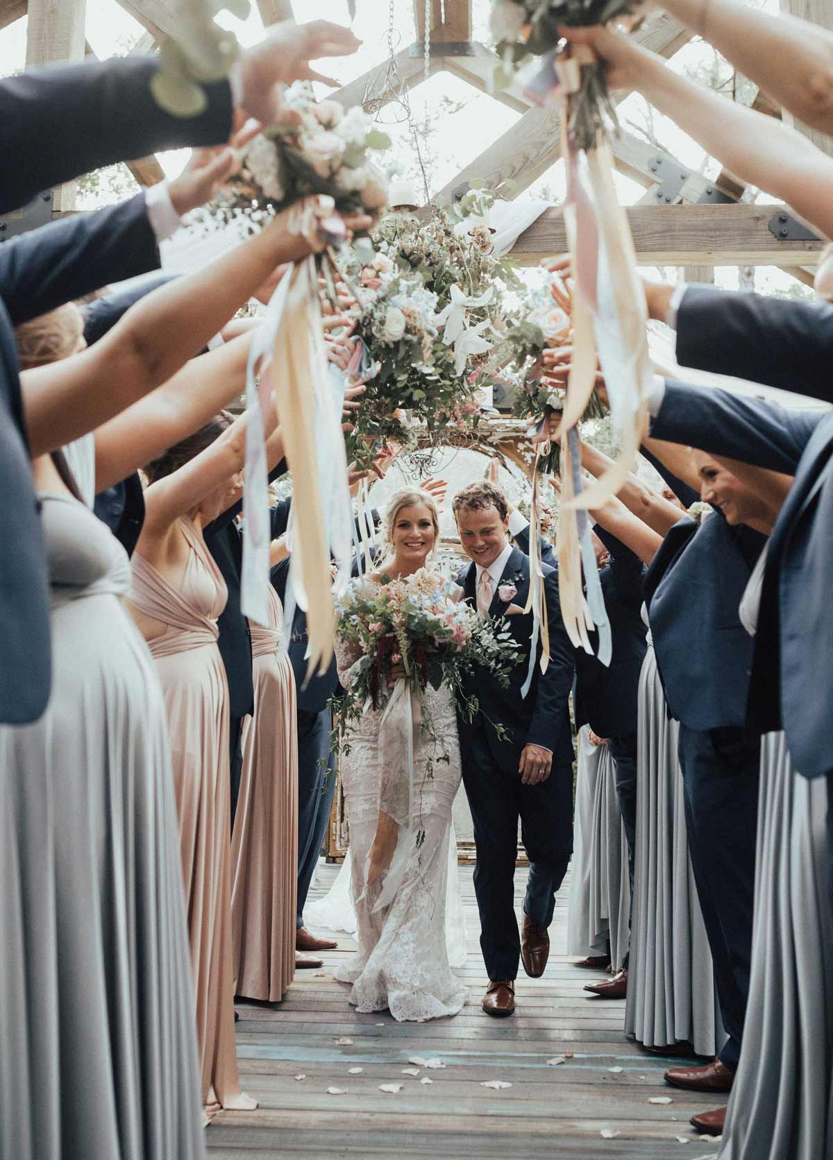 Bride and groom walk through an archway of raised bouquets held by wedding guests.