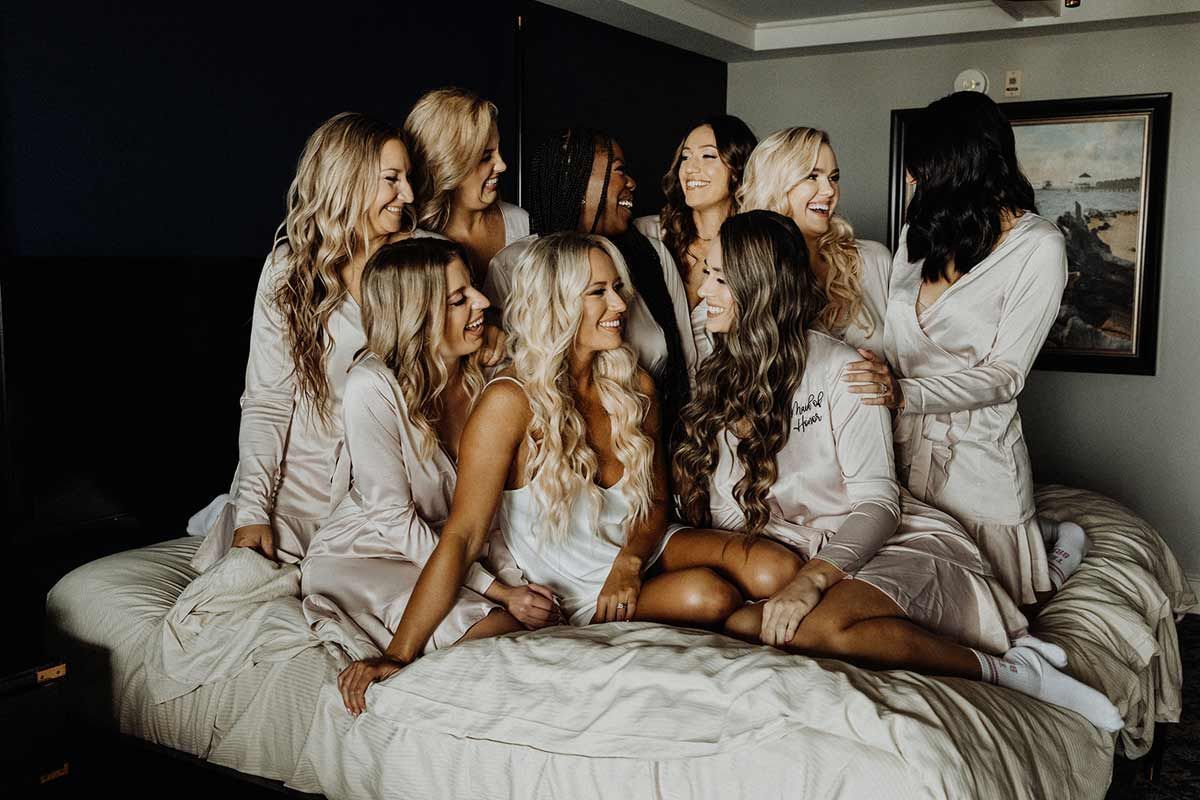Bridesmaids in robes, laughing and posing on a bed in a hotel room.